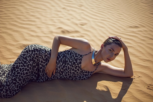 Foto brünette frau in einem langen leopardenkleid liegt mit dem rücken in der wüste bei sonnenuntergang und geht überall hin