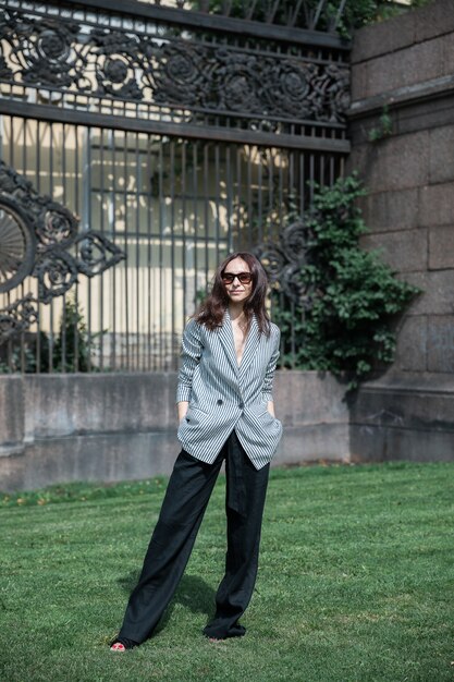 Brünette Frau in einer gestreiften Jacke und schwarzen Hosen, Sonnenbrille und Tasche String Bag.