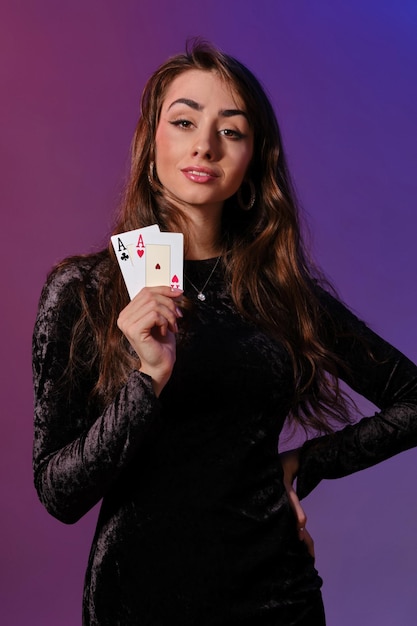 Brünette Frau in schwarzem Samtkleid zeigt zwei Spielkarten, die vor buntem Hintergrund gam posieren