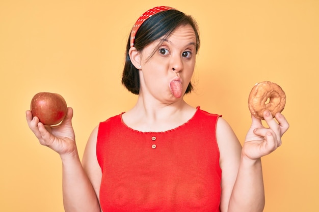 Brünette Frau mit Down-Syndrom hält roten Apfel und Donut in der Hand und streckt die Zunge heraus, glücklich und mit lustigem Gesichtsausdruck