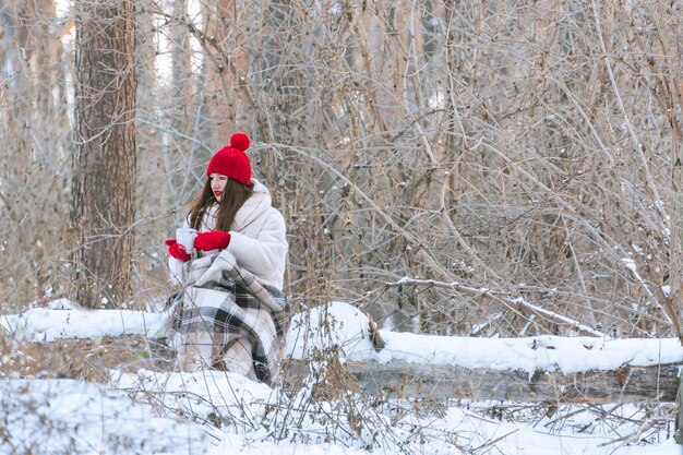 Brünette Frau mit rotem Hut und Handschuhen im Winterwald trinkt Tee. Platz kopieren.