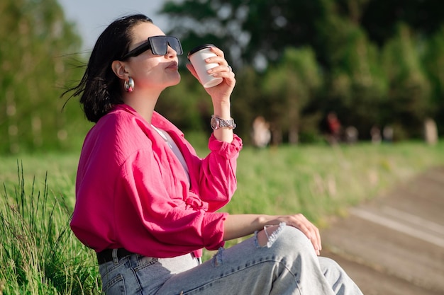 Brünette Frau mit Sonnenbrille sitzt am Seeufer und chillt in den Sonnenstrahlen im Freien