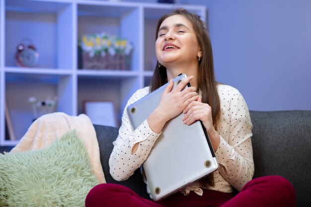 Brünette Frau umarmen Laptop, sie liebt ihre Arbeit. Glückliches junges Mädchen, Studentenumarmungsbuch, Notizbuch, liebe das Lernen, Lesen, beendete Hausaufgaben. Sitzen in ihrem Zimmer am Tisch mit Laptop