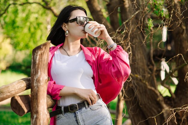 Brünette hübsche Frau mit Sonnenbrille, die eine Kaffeetasse hält und im Park spazieren geht