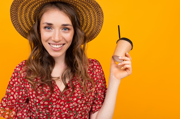 Brünette in einem Hut mit einem Glas Kaffee in der Hand auf dem Hintergrund einer gelben Wand