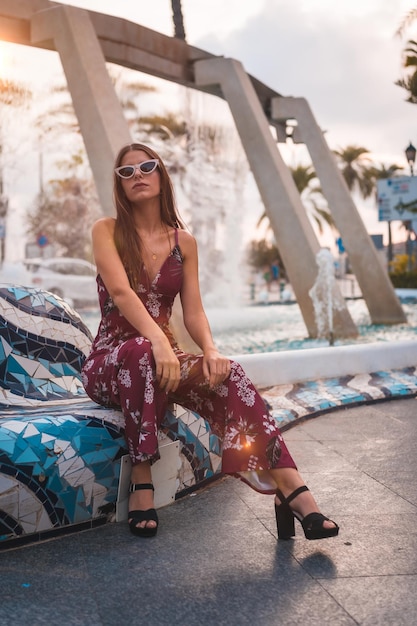 Brünette in einem kastanienbraunen Blumenkleid genießt den Sommer in der goldenen Stunde in einem Stadtbrunnen