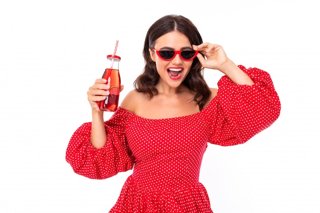 Brünette in einem roten Kleid mit einer Flasche frischem Saft tanzt auf einem weißen Hintergrund
