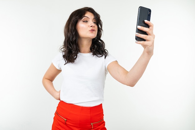 Brünette junge Frau in einem weißen T-Shirt nimmt ein Selfie auf ihrem Smartphone auf einem weißen isolierten Hintergrund mit Kopienraum.