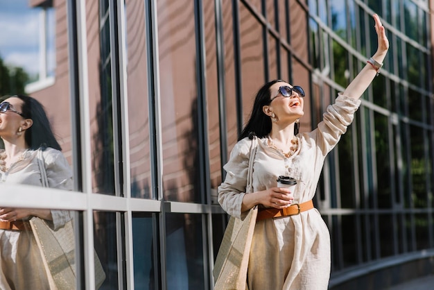 Brünette junge Frau mit Sonnenbrille und Tasche mit Kaffee zu Fuß in der Stadt. Lifestyle-Porträt der Frau