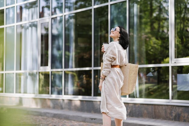 Brünette junge Frau mit Sonnenbrille und Tasche mit Kaffee zu Fuß in der Stadt. Lifestyle-Porträt der Frau