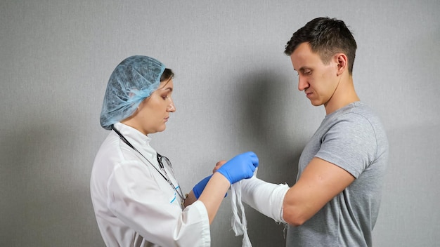 Brünette Krankenschwester in Handschuhen verwendet weißen Schwad, um den gebrochenen Unterarm eines jungen Mannes zu verbinden, der in der Klinik ein graues T-Shirt trägt.