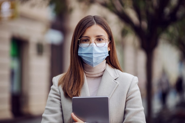 Brünette mit Gesichtsmaske, die auf der Straße steht und Tablette während des Koronavirus hält.