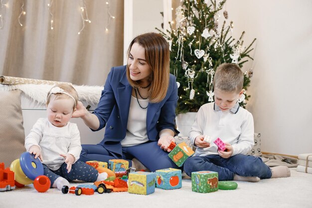 Brünette Mutter und ihre Kinder sitzen zu Hause auf dem Boden neben dem Weihnachtsbaum. Die Familie spielt mit bunten Spielsachen. Frau und Mädchen, die weiße und beige Kleidung tragen.