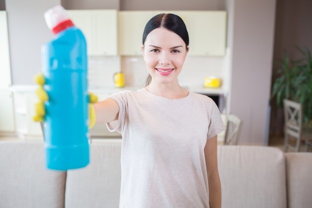 brünette steht und zeigt flasche mit blauer flüssigkeit. Frau schaut vor der Kamera und lächelt. Sie ist mitten im Raum.