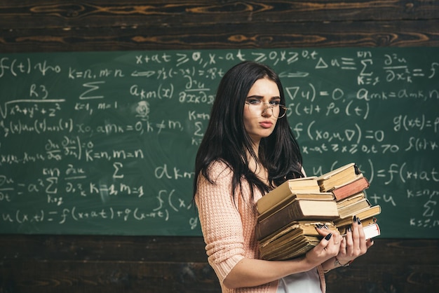 Brünette Studentin, die mit zwei Haufen Büchern vor grünem Brett voller Schrift geht. Mathe-Studium zur Prüfungsvorbereitung