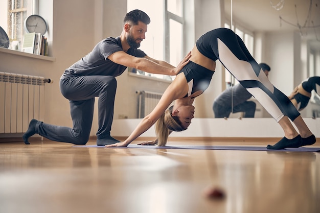 Brünette Trainerin hilft jungen blonden Frauen in Sportkleidung im Fitnessstudio und zeigt gleichzeitig die richtige Pose