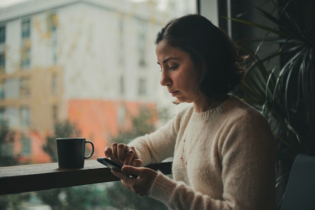 Brünette türkische Frau, die ihr Handy ansieht, während sie in einem Café einen Kaffee trinkt, ist sie in ernster Stimmung