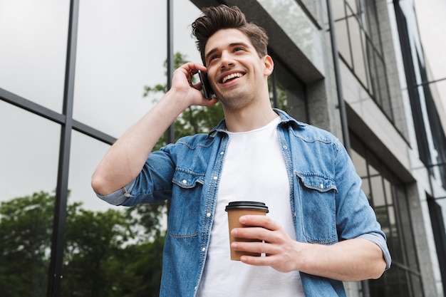 Brünetter junger Mann in Freizeitkleidung, der Kaffee zum Mitnehmen trinkt und am Handy spricht, während er über dem Gebäude steht