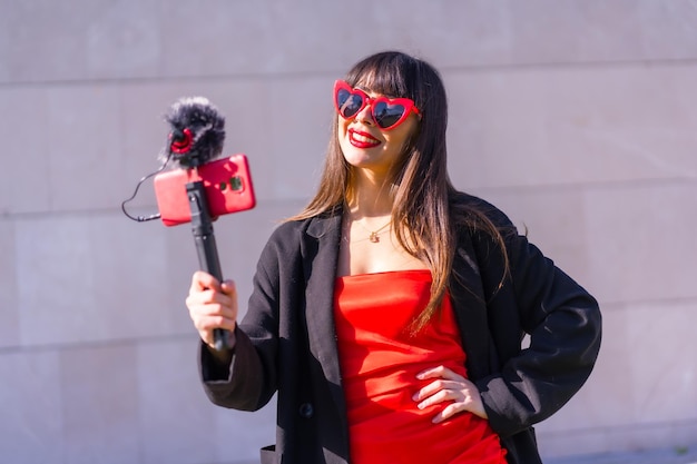 Brünettes kaukasisches Mädchen, das am Valentinstag einen Vlog für soziale Netzwerke macht, Sonnenbrille mit roten Herzen, Telefon und Mikrofon auf grauem Hintergrund