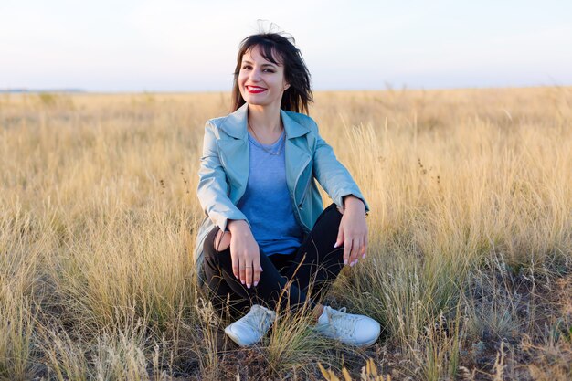 Brünettes Mädchen in einer blauen Jacke liegt in einem Feld auf gelbem Gras y