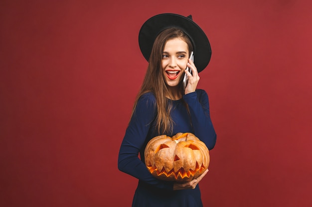 Bruja de Halloween con una calabaza tallada - aislada sobre fondo rojo. Mujer joven emocional en disfraces de Halloween. Usando el teléfono.