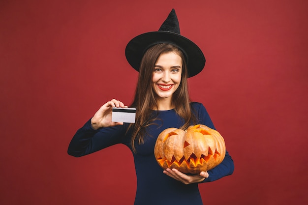 Bruja de Halloween con una calabaza tallada y tarjeta de crédito - aislada sobre fondo rojo. Mujer joven emocional en disfraces de Halloween.