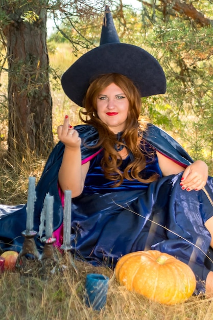 Una bruja pelirroja con una capa azul y un sombrero en el bosque con una calabaza en las manos evoca velas