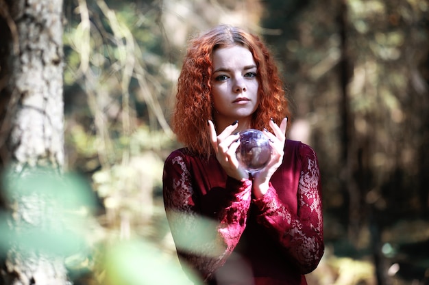 La bruja pelirroja realiza un ritual con una bola de cristal en el bosque.