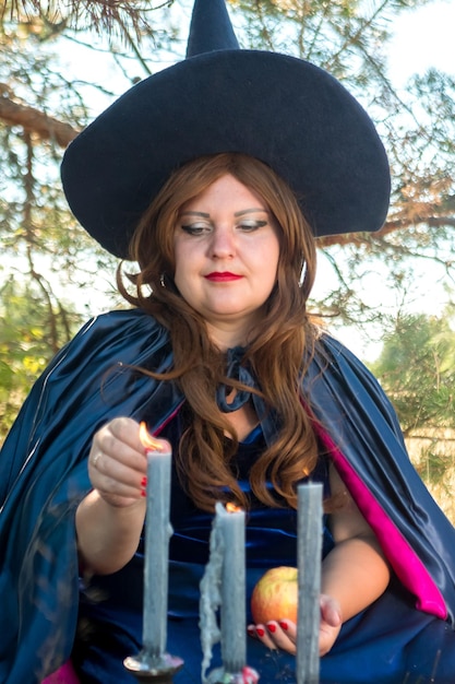 Una bruja pelirroja con un sombrero puntiagudo y una capa enciende velas en un viejo candelabro en el bosque