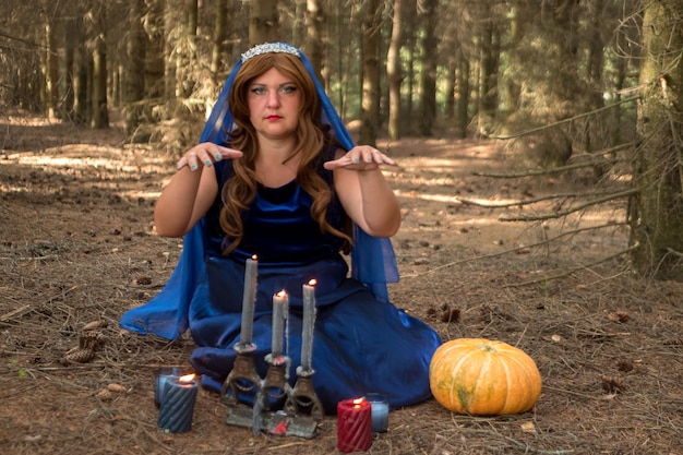 Una bruja pelirroja con un vestido azul y un velo dice fortunas en el bosque con velas en un viejo candelabro