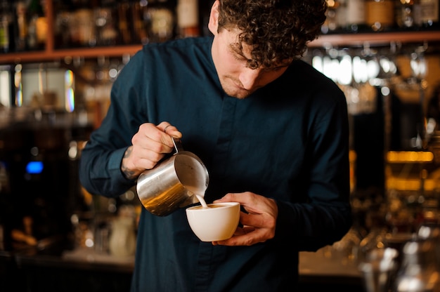 Brunet Barkeeper gießt eine Milch aus dem Stahlkessel in die weiße Keramik Tasse Kaffee