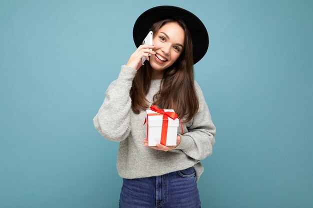 Brunet Frau lokalisiert über blaue Hintergrundwand, die schwarzen Hut und grauen Pullover hält Geschenkbox hält, die auf Handy spricht und Kamera betrachtet.