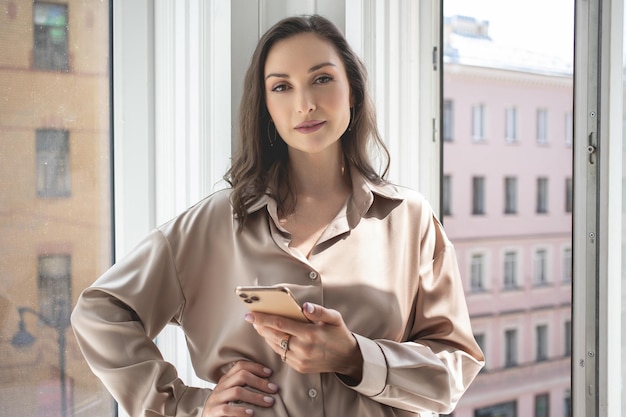 Foto bruneta elegante em uma camisa solta retrato junto à janela luz solar brilhante uma mulher usando um telefone