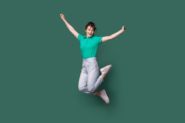 Brunette kaukasische Frau in Jeans und T-Shirt, die auf eine grüne Studiowand springen und lächeln