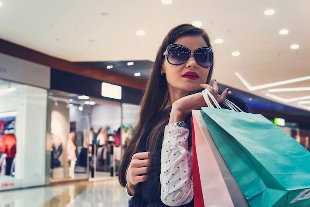 Brunette Mädchen in dunklen Gläsern halten Haufen von Einkaufstüten im Mall-Hintergrund Junges attraktives Mädchen mit rotem Lippenstift nach dem Kauf im Laden Nebenkosten der Frau Weiblicher Spaziergang für den Kauf von Dingen