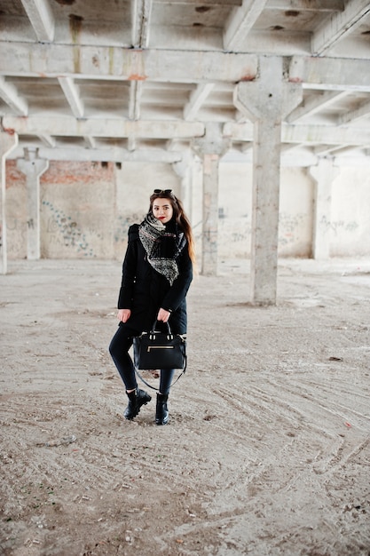 Brunette stilvolles lässiges Mädchen in Schal und Handtasche gegen verlassenen Fabrikplatz.