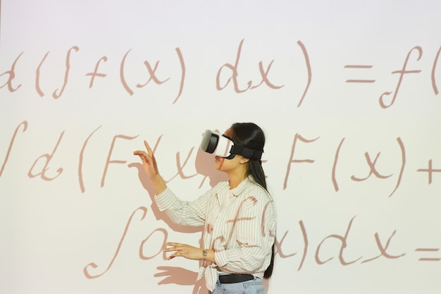 Brunette Studentin in Virtual-Reality-Headset zeigt auf Projektionsfläche während der Arbeit mit mathematischen Gleichung