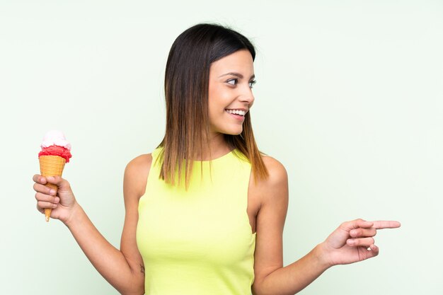 Brunettefrau, die eine Kornetteiscreme über der grünen Wand zeigt auf die Seite hält, um ein Produkt darzustellen