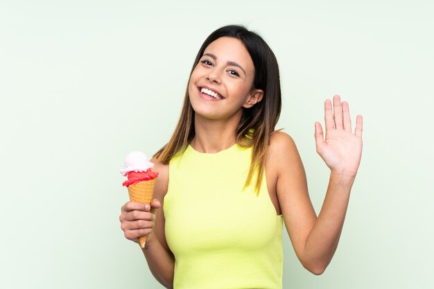 Brunettefrau, die eine Kornetteiscreme über lokalisierter grüner Wand begrüßt mit der Hand mit glücklichem Ausdruck hält