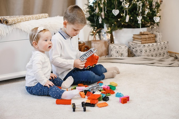 Brunettejunge und -mädchen, die zu Hause nahe Weihnachtsbaum sitzen. Geschwister spielen zusammen. Jungen und Mädchen, die blaue und beige Kleidung tragen.