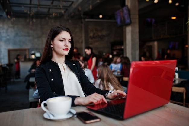 Brunettemädchen, das auf Café mit Schale Cappuccino, arbeitend mit rotem Laptop sitzt.