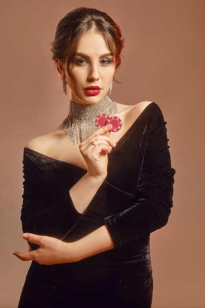 Brunettemodell im schwarzen Kleid und im glänzenden Schmuck. Sie zeigt zwei rote Chips und posiert vor braunem Studiohintergrund. Schürhaken, Kasino. Nahansicht
