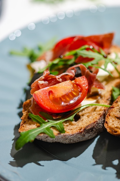 Bruschetta com baguete escura, jamon, rúcula e tomate em uma mesa de azulejos brancos, luz solar forte e brilhante. Um lanche saudável e elegante.