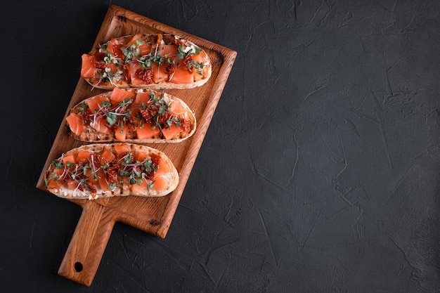 Bruschetta com salmão, tomate seco, ervas, queijo cottage, sobre fundo preto, placa de madeira