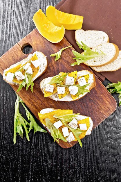 Bruschetta mit gebackenem Kürbis, gesalzenem Feta-Käse, Ricotta, Rucola und Gewürzen, Handtuch- und Gemüsescheiben auf Holzbretthintergrund von oben