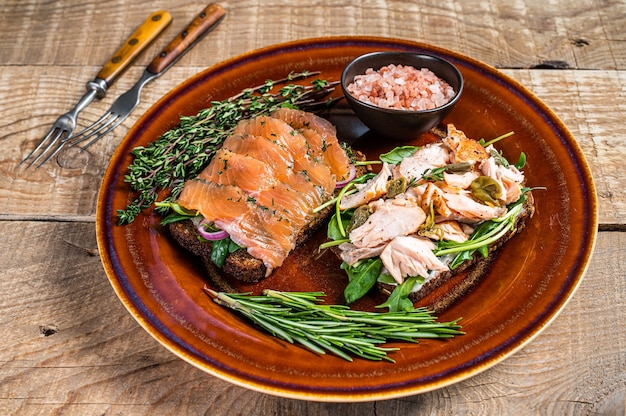 Bruschetta mit warmem und kaltem Räucherlachs, Rucola, Kapern auf einem rustikalen Teller mit Kräutern. hölzerner Hintergrund. Ansicht von oben.