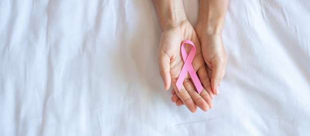 Brustkrebs-Bewusstseinsmonat Oktober, Hand der erwachsenen Frau, die Pink Ribbon auf rosafarbenem Hintergrund hält, um Menschen zu unterstützen, die leben und krank sind. Konzept des Internationalen Frauen-, Mutter- und Weltkrebstages