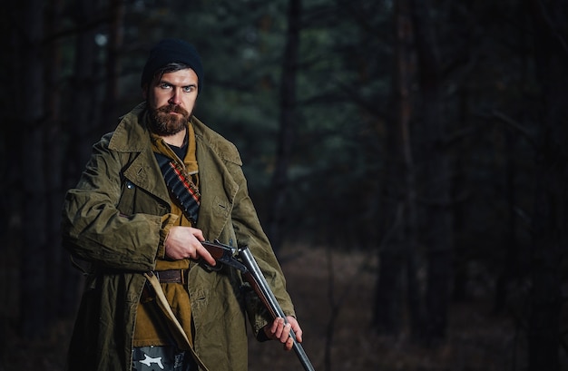 Foto un brutal cazador barbudo con un sombrero oscuro y una chaqueta de color caqui en una capa larga sostiene una pistola descargada contra el fondo de un bosque oscuro