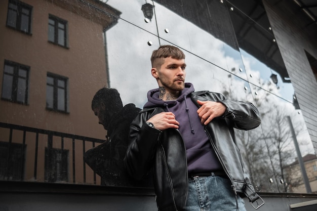 Brutal guapo hipster en ropa casual de moda usa chaqueta de cuero de moda rock cerca de un edificio moderno negro en la calle