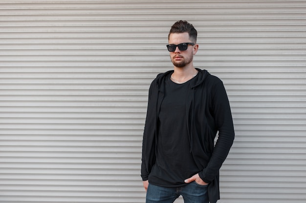 Brutal guapo joven inconformista con barba en una elegante camiseta con una sudadera con capucha negra de moda en gafas de sol en jeans de moda se encuentra cerca de una pared de metal vintage